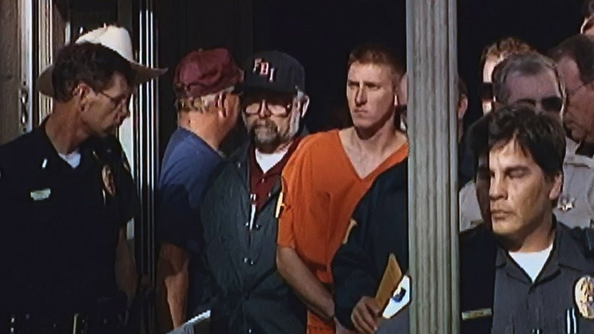 Timothy McVeigh in an orange jumpsuit surrounded by plainsclothed police officers.