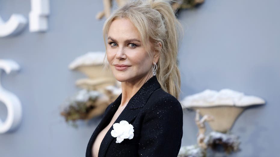 Nicole Kidman smiling, wearing a black blazer with a white flower pinned to the lapel
