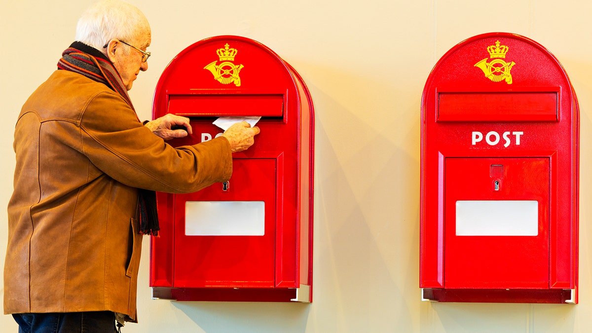 An elderly man wearing a brown jacket and a scarf around his neck puts a letter in one of two Danish mailboxes pictured next to each other.