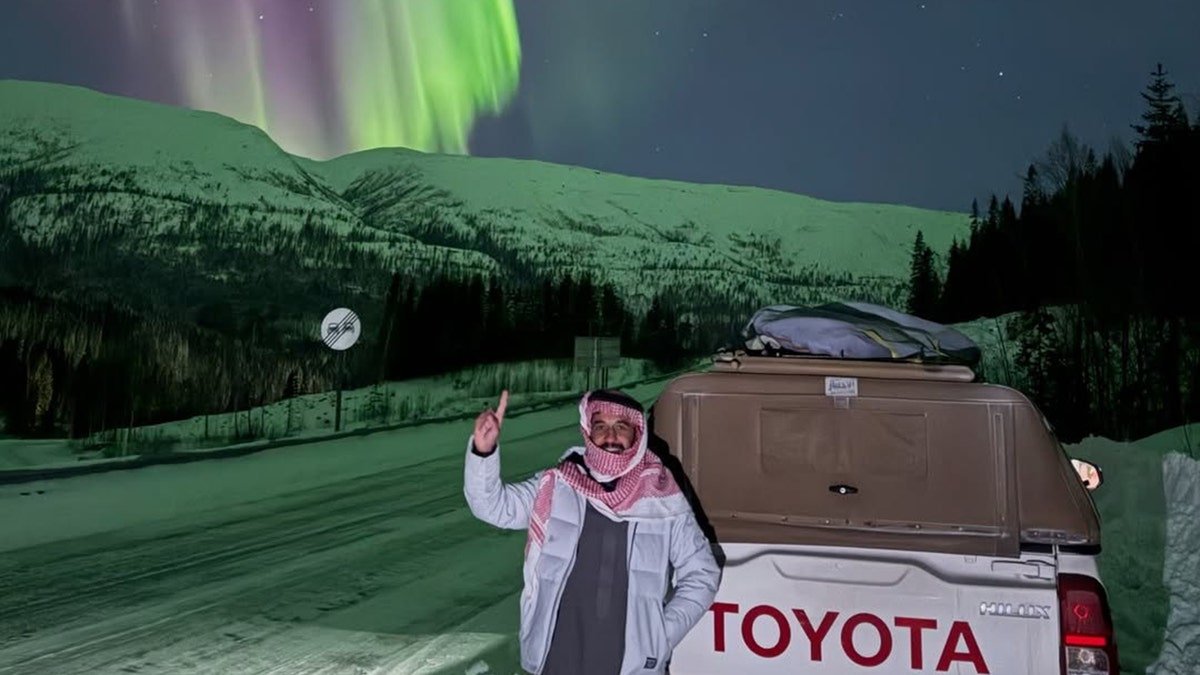 Malik Al-sultan standing beside a Toyota pickup truck under the northern lights on a snowy mountain road at night.