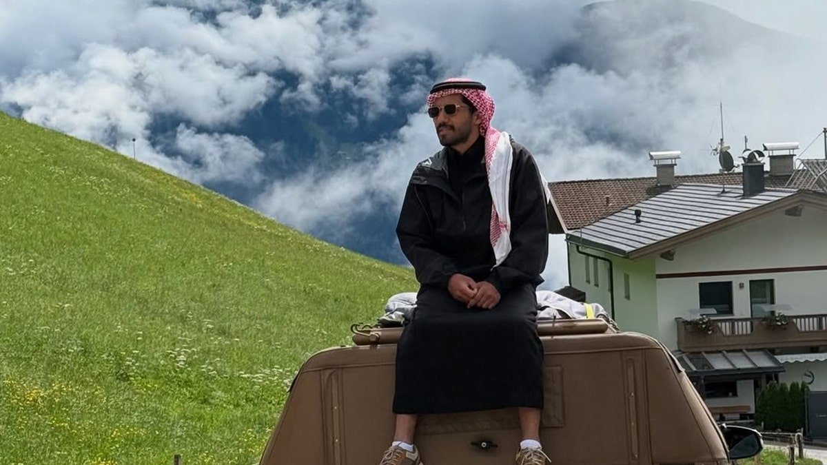 Malik Al-sultan sitting on top of a Toyota pickup truck in a green alpine village with mountains and clouds.