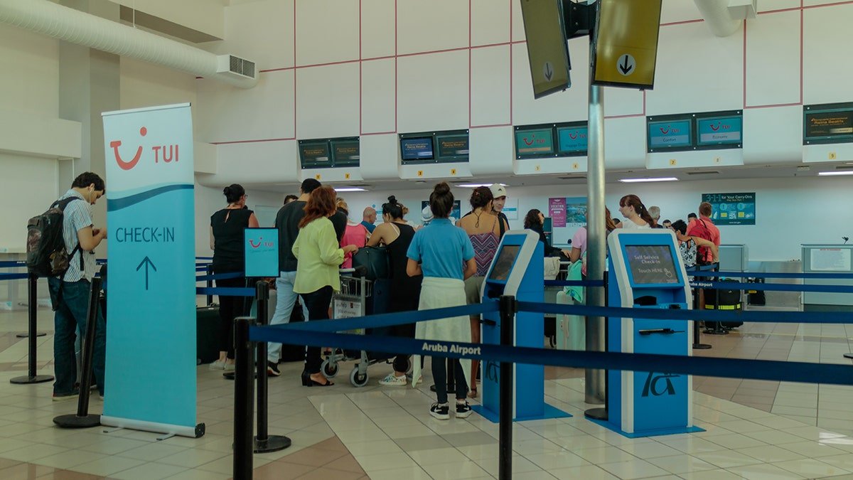 Travelers waiting in line at check-in counters inside Aruba Airport, with self-service kiosks and airline signage visible.