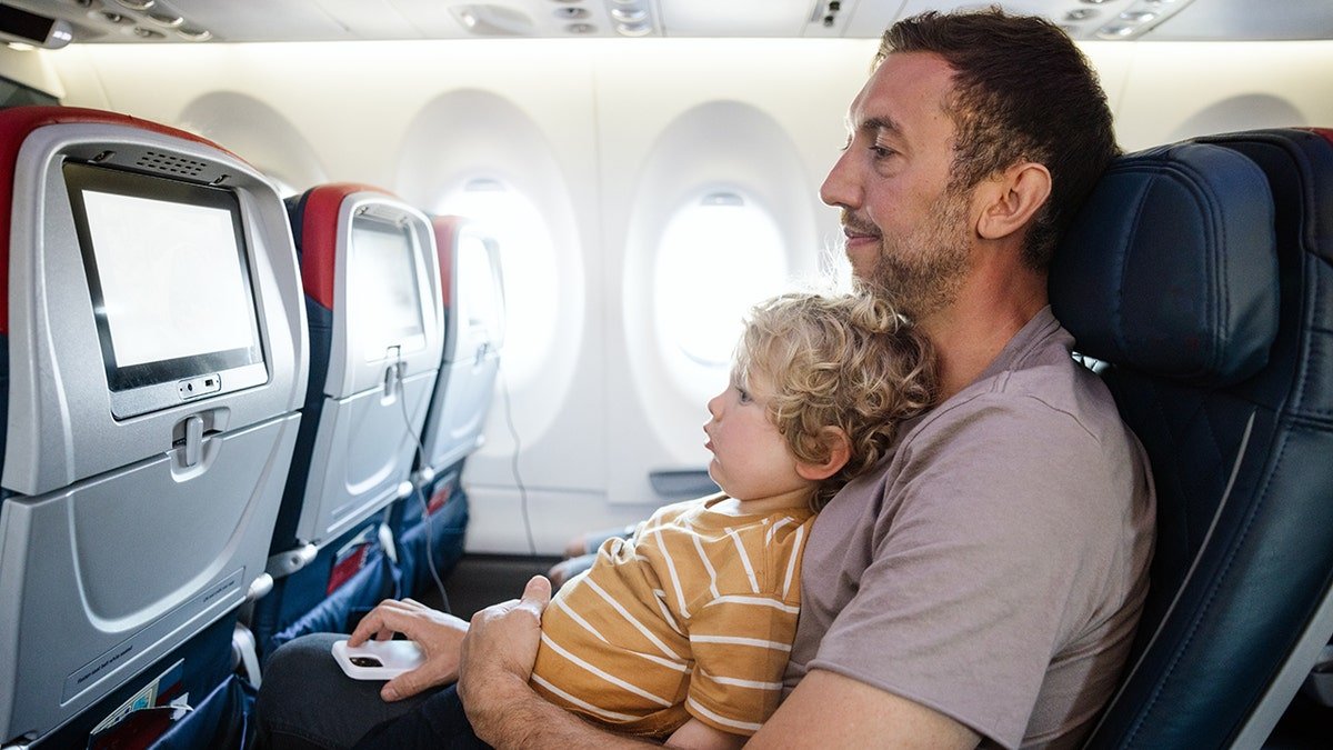 Father and young son sitting together on airplane watching in-flight entertainment screen.