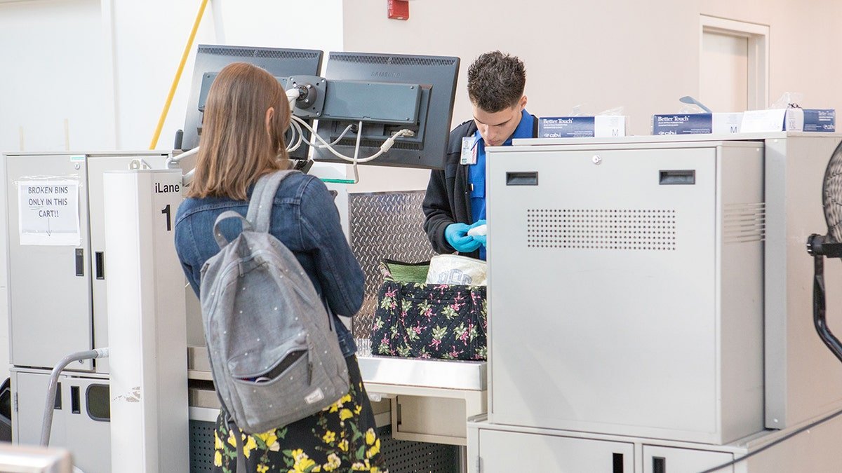 Airport security officer wearing blue gloves inspects a passenger’s floral bag at a TSA screening checkpoint.