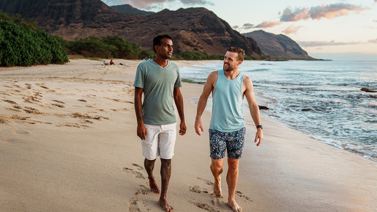 Two men walking barefoot along a sandy beach with mountains and ocean waves at sunset.
