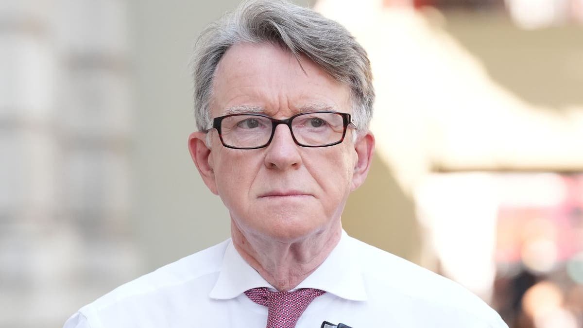 Peter Mandelson walks outdoors carrying a folder while wearing glasses and a red tie