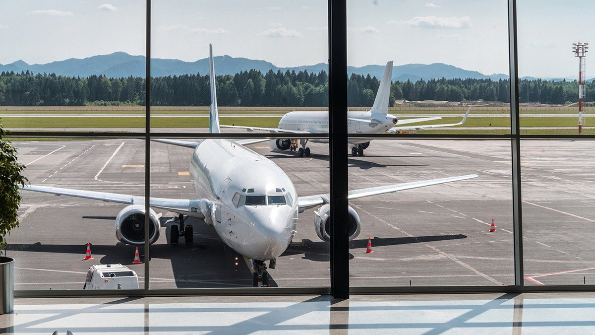 Commercial passenger airplane parked at an airport gate, viewed through terminal windows with runway and mountains in the background.