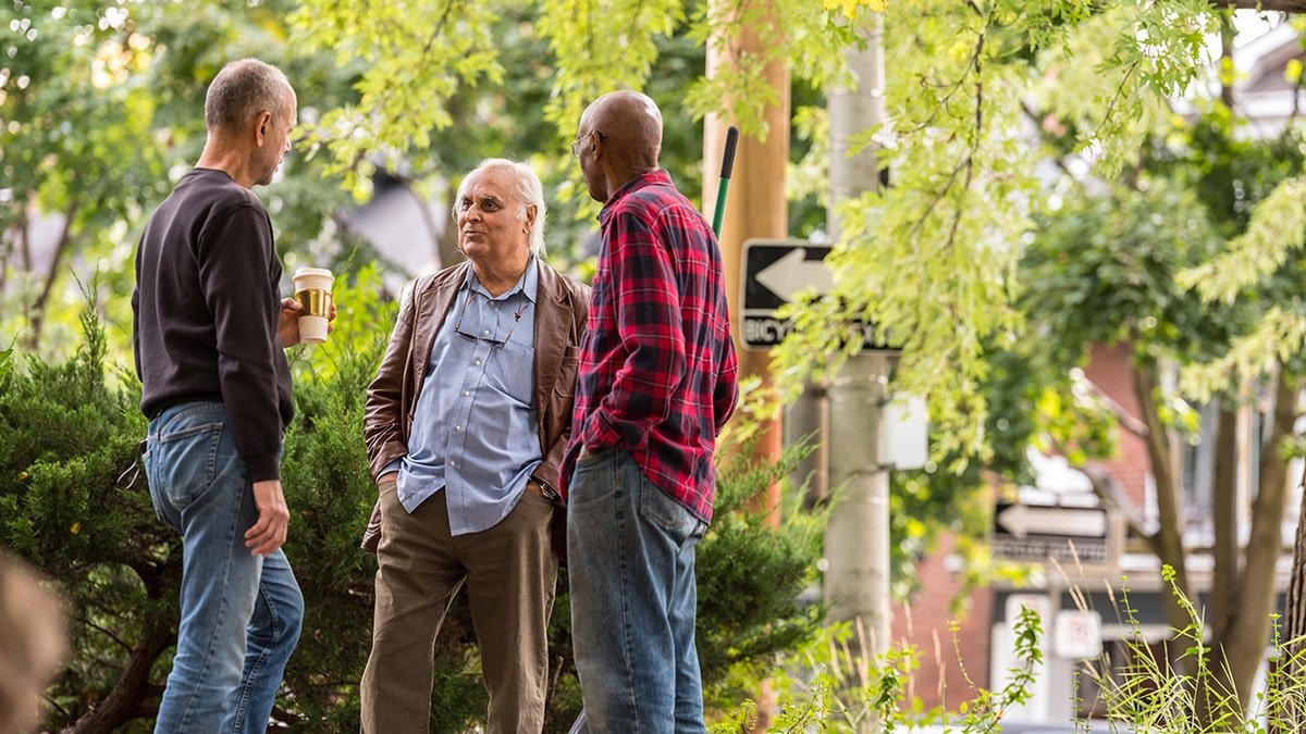 Three older men chatting outdoors in a leafy neighborhood, one holding a coffee cup, standing near a sidewalk and greenery.