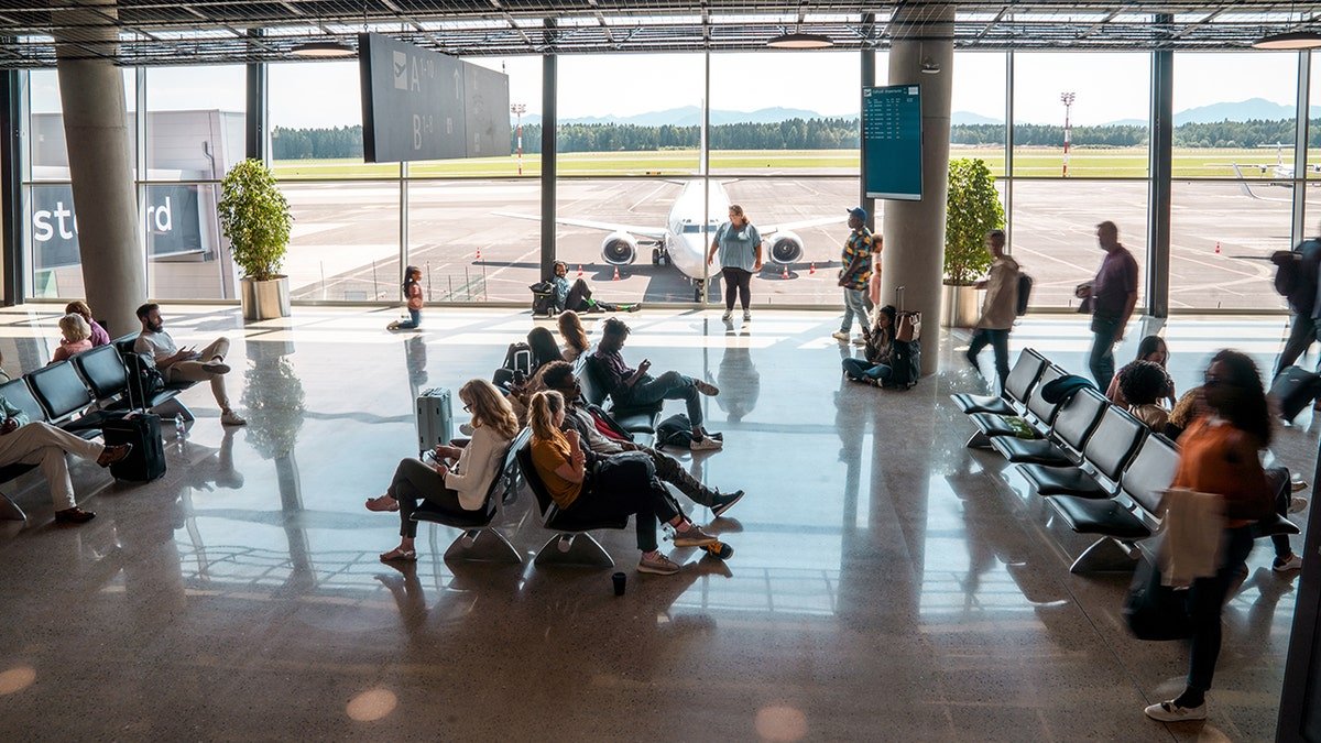 Passengers waiting in an airport terminal with a commercial airplane visible through large windows at the gate.