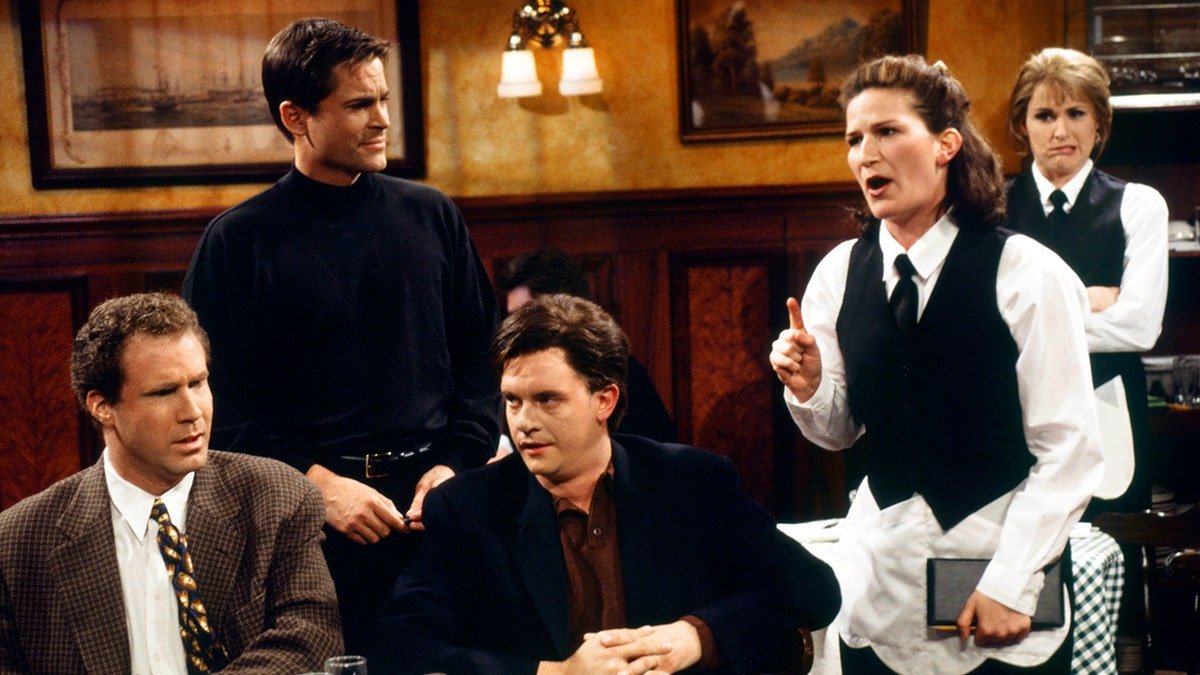 Cast members in a restaurant scene from Saturday Night Live (SNL), with a waitress pointing emphatically while two men sit at a table and two others stand behind them.