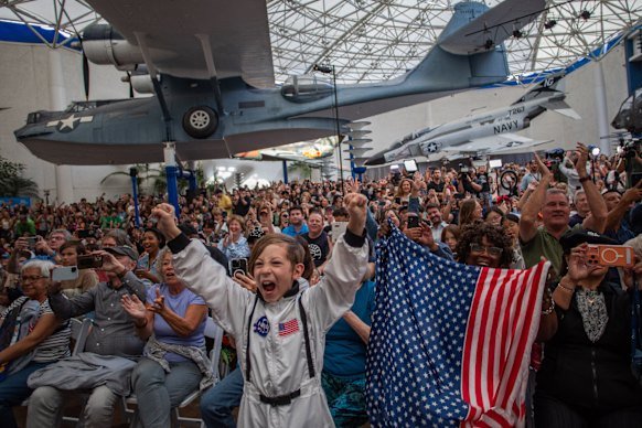 A young boy wears an astronaut costume next to a woman waving a flag as they watch a live broadcast of the return of the Artemis II crew.
