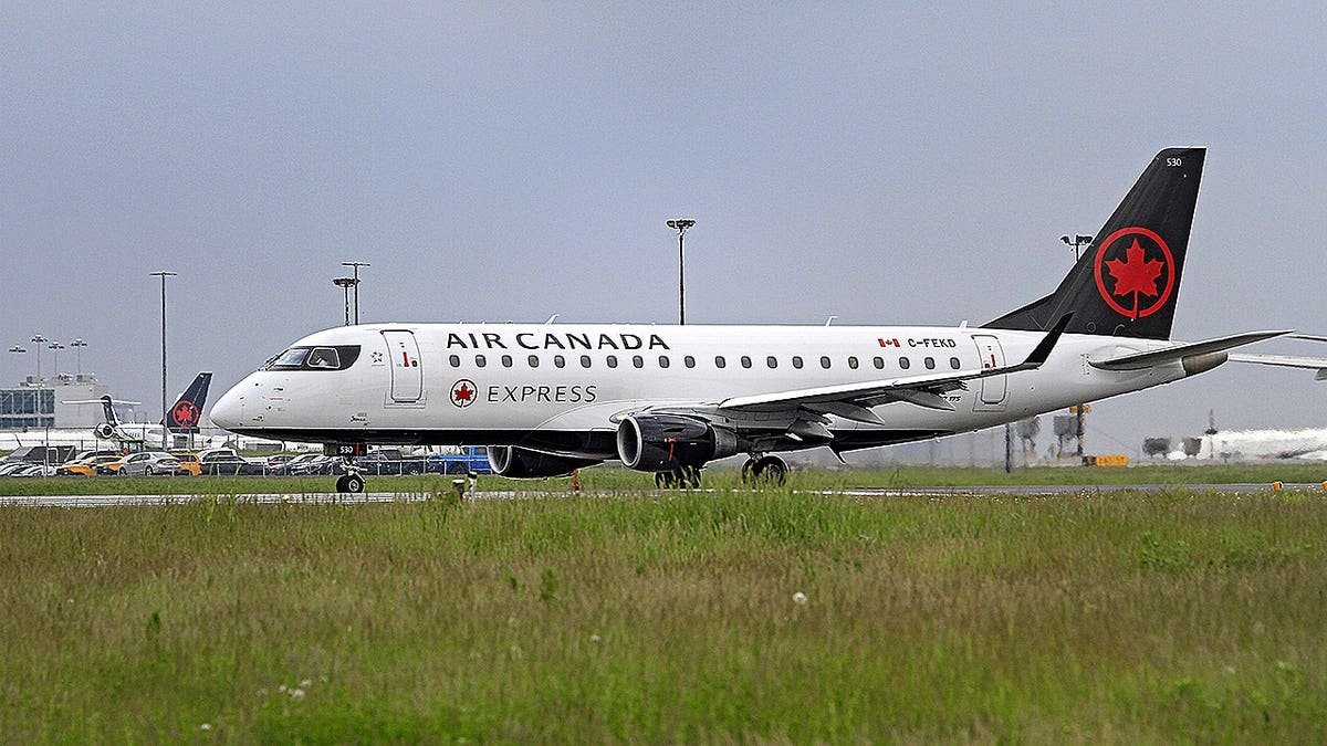 Air Canada Express jet taxis on a runway at an airport, with terminal buildings and other aircraft in the background.