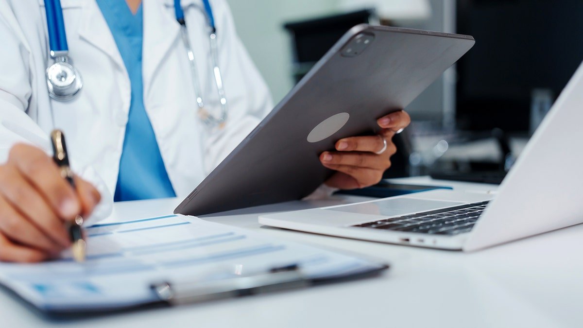 A doctor looking at a tablet and writing on a clipboard.
