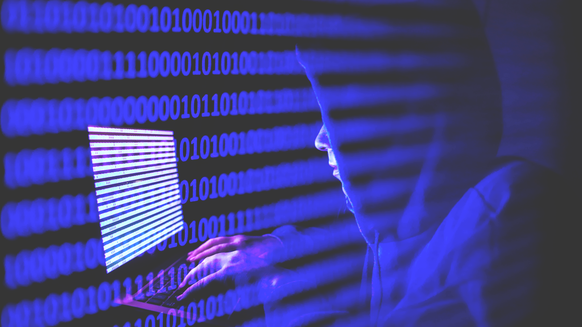 A person dressed as an internet hacker working on a laptop with binary code on the screen.