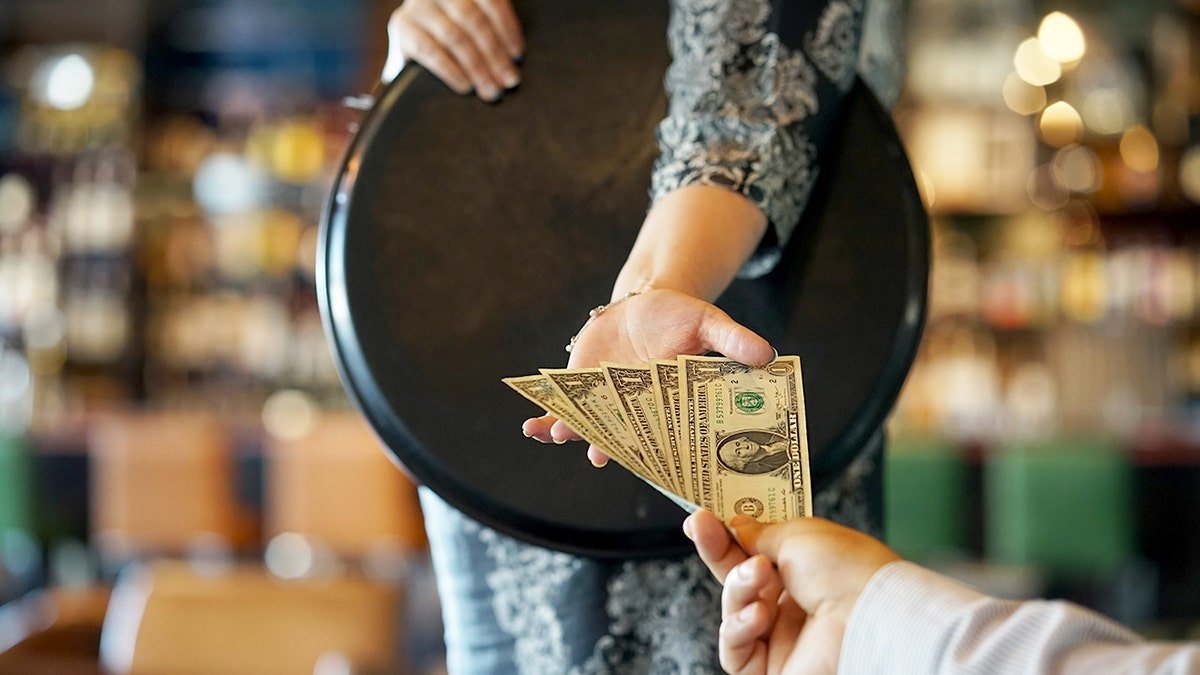 A waitress hand holding a serving tray receiving a five dollar cash tip from a customer