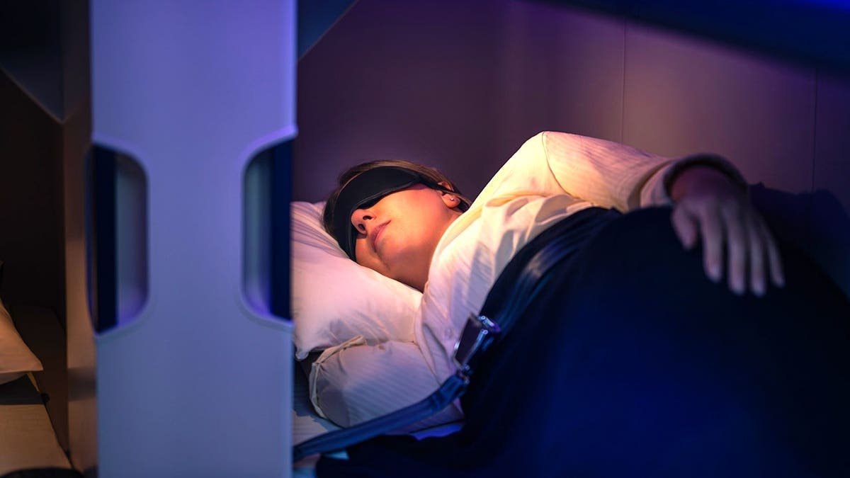 Passenger sleeping in Air New Zealand Economy Skynest pod with lie-flat bed and sleep mask on ultra-long-haul flight.
