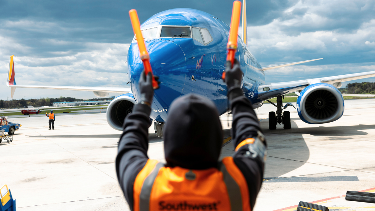 Ground crew member in a safety vest signals with orange wands to guide a Southwest Airlines plane on the airport tarmac under cloudy skies.