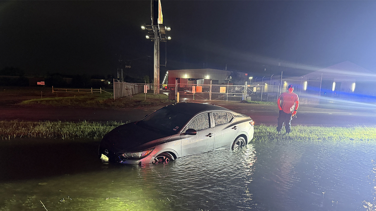 Person stands next to partially submerged car in floodwaters during Texas storm at night.