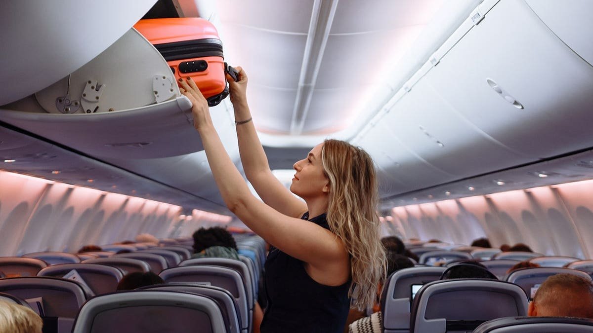 Woman placing pink hard-shell suitcase in airplane overhead storage compartment