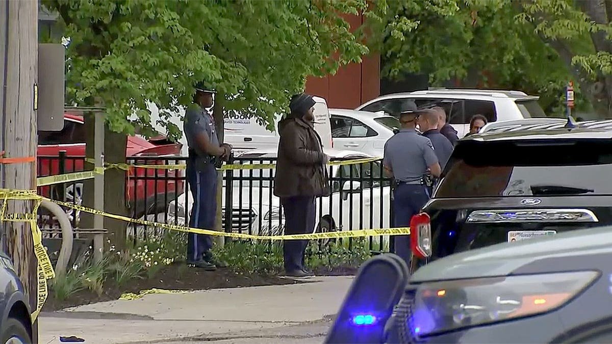 Police officers stand near yellow crime scene tape along a sidewalk in Cambridge, Massachusetts, as vehicles and bystanders are visible nearby.