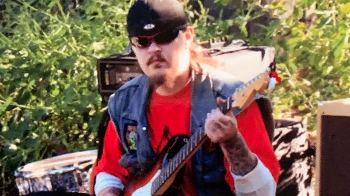 Keith Palumbo wearing a red shirt and a denim vest playing guitar outdoors.