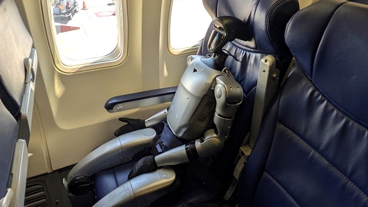 Humanoid robot seated in a window seat on an airplane, secured with a seatbelt.