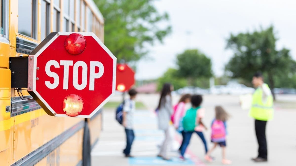 Children with backpacks cross a street guided by a crossing guard near a yellow school bus with its stop sign extended.
