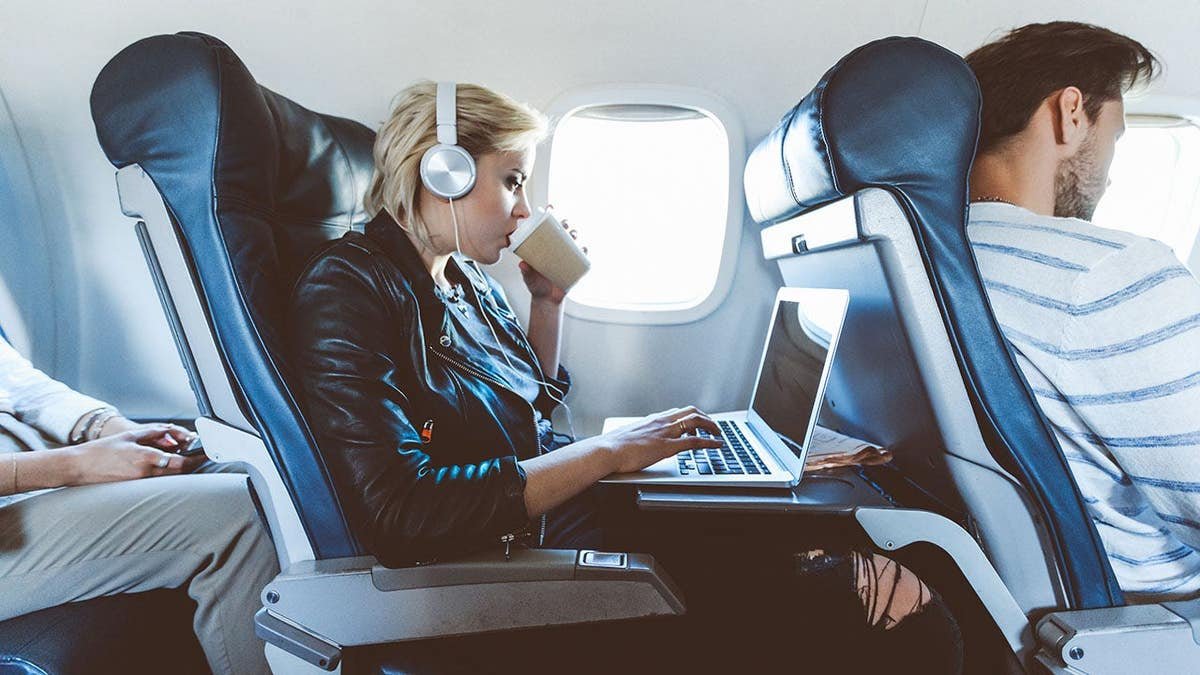 Young blonde woman sitting inside an airplane using a laptop and drinking coffee