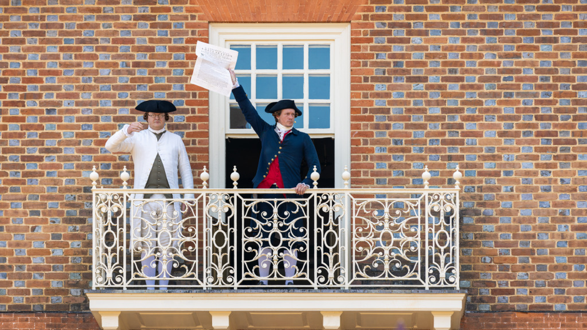 A man holds up a paper on a balcony, wearing colonial clothes.