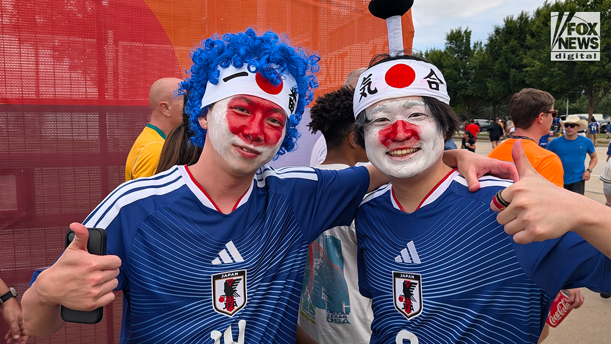 Two Japan soccer fans wearing blue national team jerseys, face paint in the colors of the Japanese flag, and headbands pose together with thumbs up outside a FIFA World Cup venue.