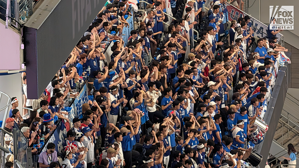 Japan national team supporters dressed in blue cheer and raise their arms from a packed stadium section during a FIFA World Cup match.