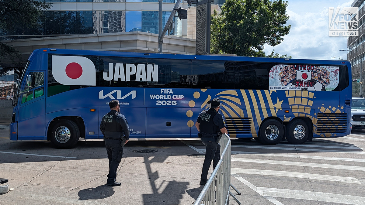 A blue FIFA World Cup 2026 team bus displaying Japan's name and flag is parked on a city street as police officers stand nearby.