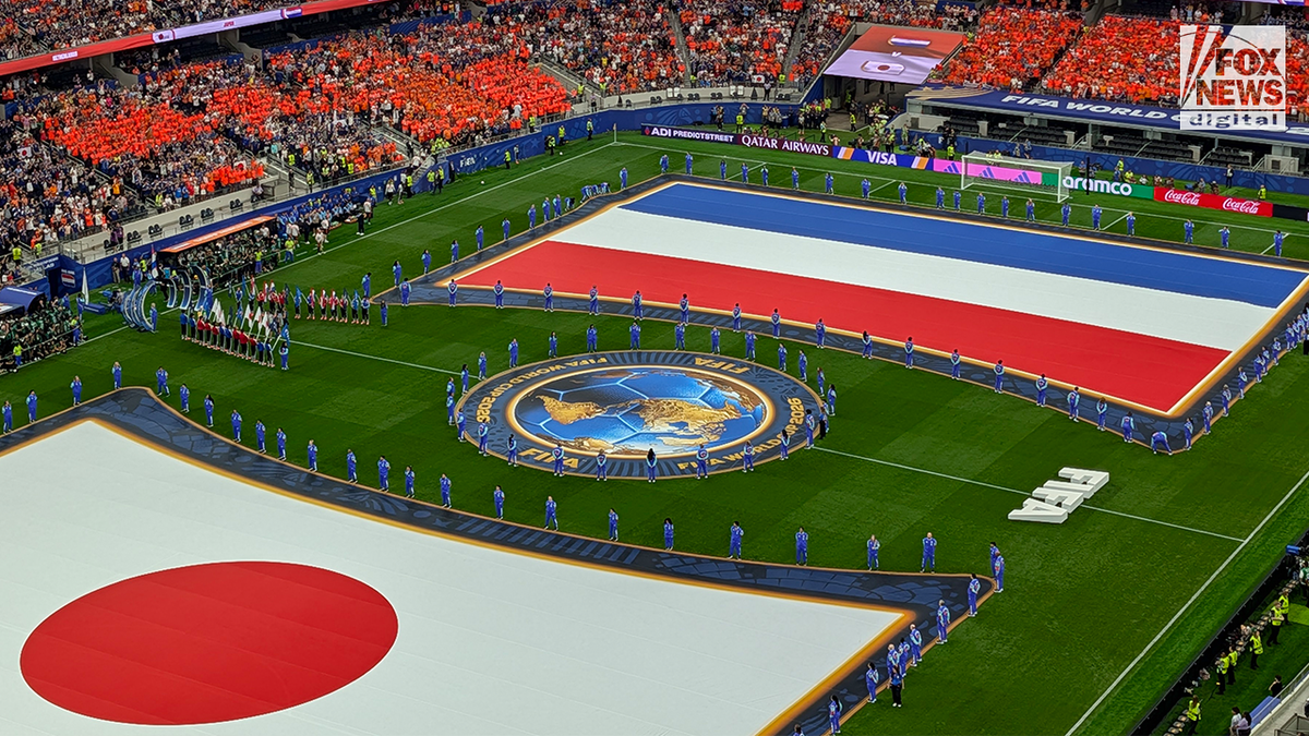 Large Japanese and Netherlands national flags are displayed across the field during a FIFA World Cup pre-match ceremony as teams and officials line the pitch in a packed stadium.