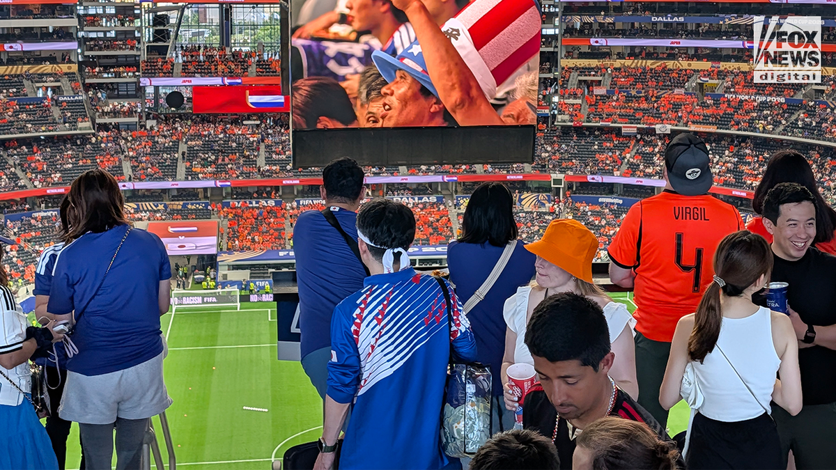 Fans wearing Japan and Netherlands team apparel watch the field from a stadium suite during a FIFA World Cup match, with a large video board visible above the stands.