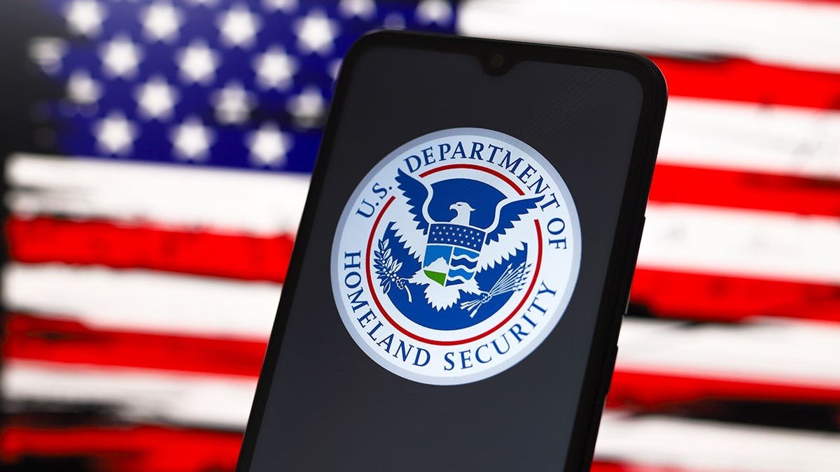 Department of Homeland Security logo on smartphone screen with American flag design on computer screen in background.