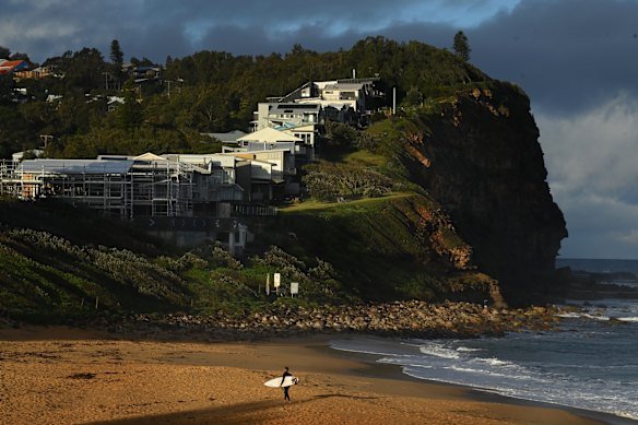 A surfer walks along Copacabana Beach under a row of luxury homes.
