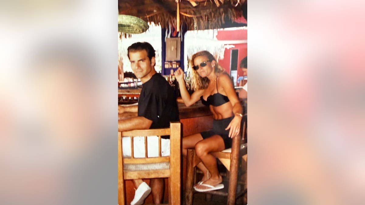 Nicole Brown Simpson in a black bikini sitting next to Joseph Perrulli in a black shirt.