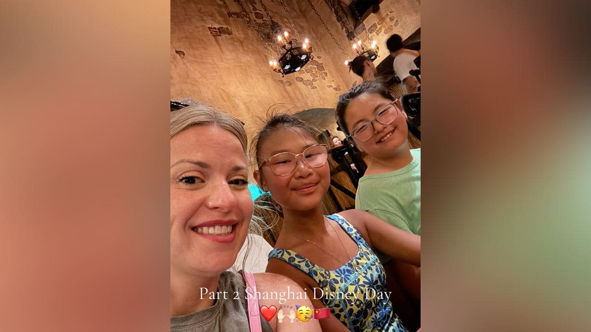A woman takes a selfie with two young girls inside a dimly lit, stone-walled space with hanging chandeliers, with the overlaid text