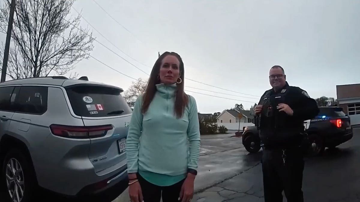 Woman in teal jacket stands in parking lot next to police officer and patrol vehicles with lights on.