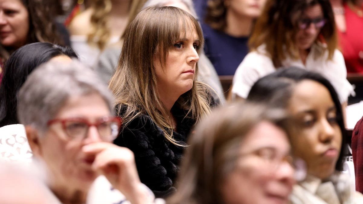 Woman with long straight hair and bangs, wearing a black textured jacket, sits among a crowd of other attendees indoors.
