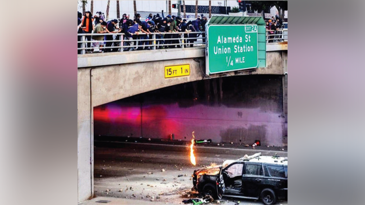 People gather on a Los Angeles freeway overpass as a California Highway Patrol vehicle burns below during anti-ICE riots.