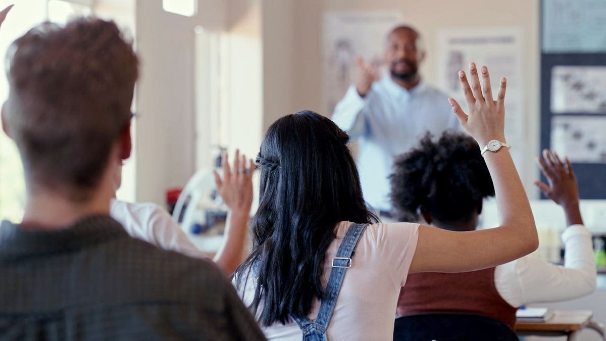 File photo showing students raising their hands in a classroom while a teacher stands at the front of the room.