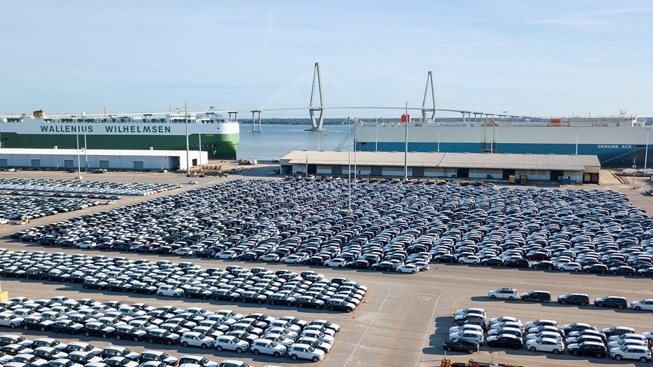 Vehicles await export at the Port of Charleston in Charleston, S.C., with cargo ships and the Arthur Ravenel Jr. Bridge in the background.