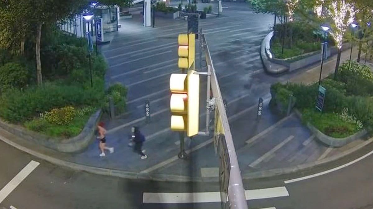 Wide surveillance view of two people running across a city intersection near a traffic signal at dusk.
