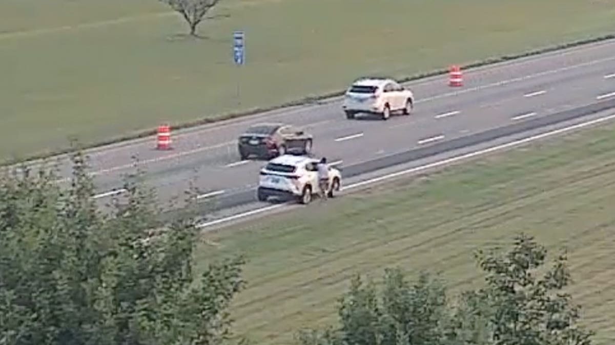 Highway camera view of a person beside a white SUV on the shoulder of an interstate near orange construction barrels.