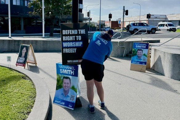 A Brethren member in shorts and a Liberal Party t-shirt puts up a poster endorsed by Morgan C Jonas.