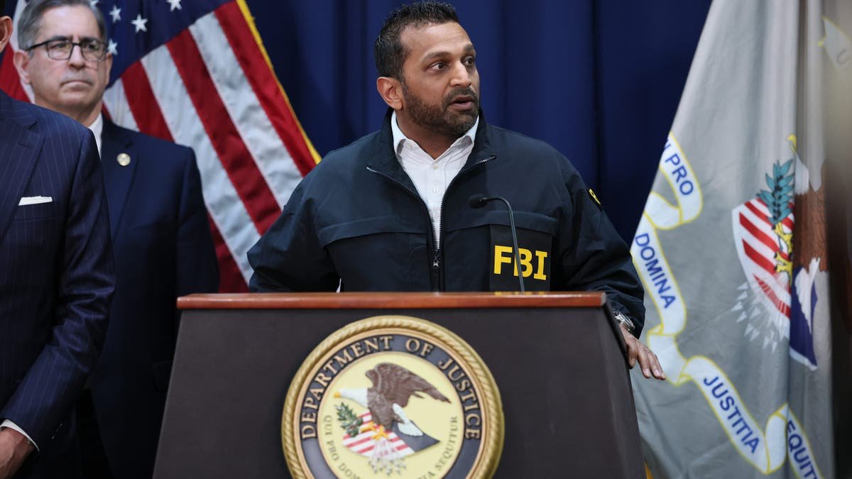 kash patel wears an black FBI jacket while standing at a DOJ podium