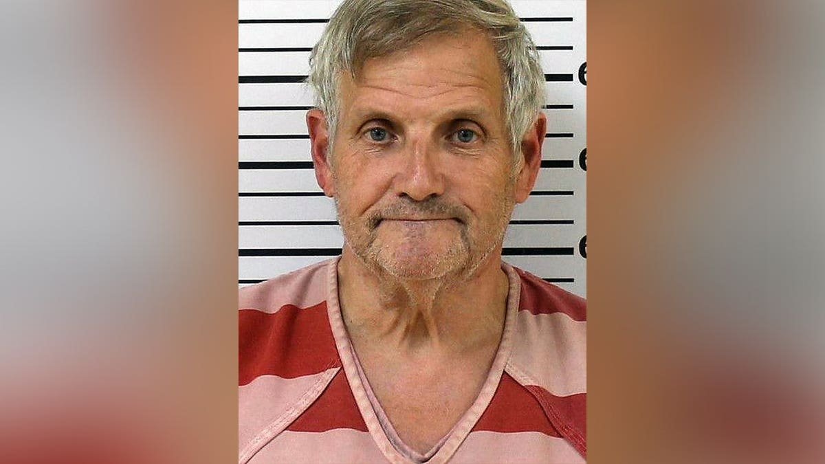 Booking photo of John Herb Hayes III, a gray-haired man wearing a red-and-pink striped shirt in front of a height chart.