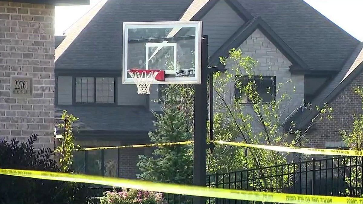 Yellow police tape stretches across the front yard of a large brick home with a basketball hoop.