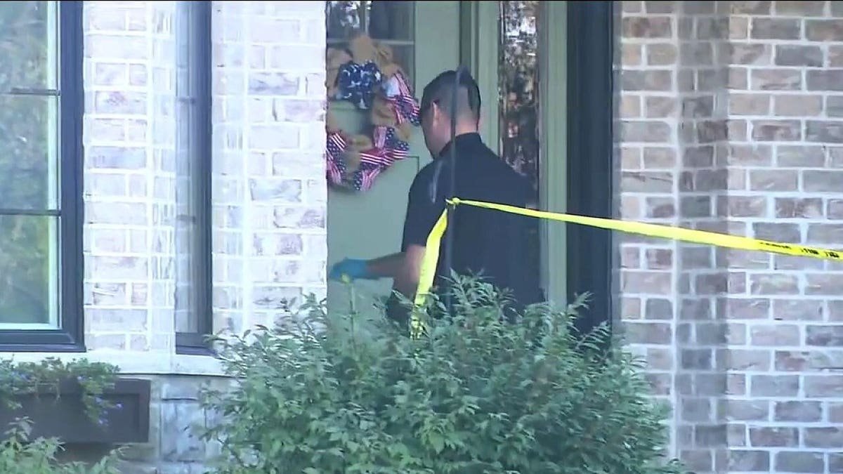 An investigator wearing sunglasses and gloves stands behind yellow police tape outside a brick home.